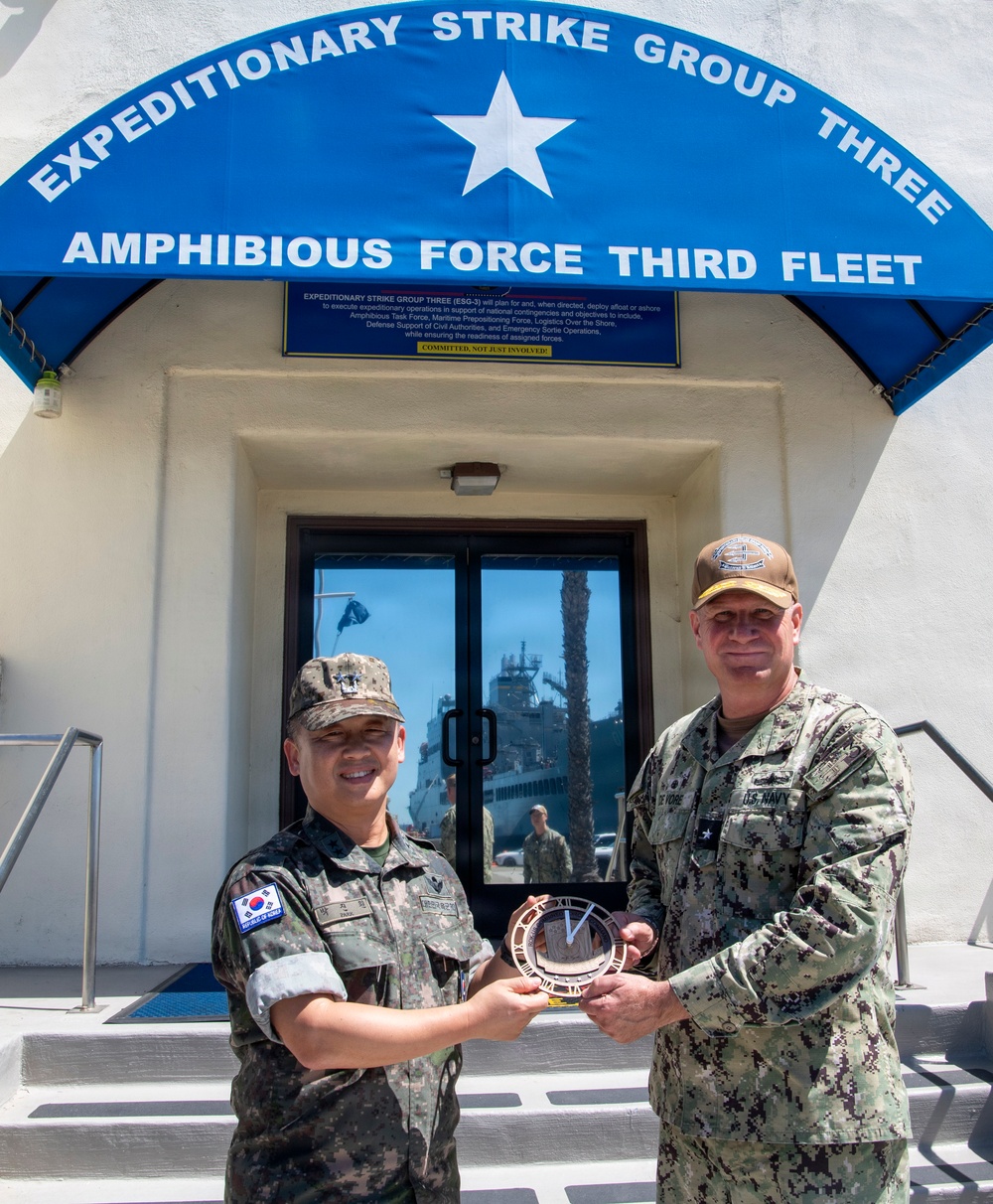Two military officers are posing for a photo in front of the Expeditionary Strike Group Three / Amphibious Force Third Fleet headquarters building. They are jointly holding what appears to be a commemorative plate or coin exchange item, suggesting an official military visit or ceremony. One officer wears a Republic of Korea (ROK) military uniform and the other wears a U.S. Navy uniform.