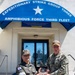 Two military officers are posing for a photo in front of the Expeditionary Strike Group Three / Amphibious Force Third Fleet headquarters building. They are jointly holding what appears to be a commemorative plate or coin exchange item, suggesting an official military visit or ceremony. One officer wears a Republic of Korea (ROK) military uniform and the other wears a U.S. Navy uniform.
