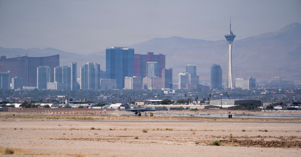 An aerial-ground level view of the Las Vegas skyline photographed from what appears to be a desert airfield or airport tarmac area. The iconic Stratosphere Tower (now known as STRAT Hotel) is prominently visible on the right side of the skyline, with numerous casino and hotel high-rises visible in the background. A military or civilian jet aircraft is visible on the runway/taxiway in the mid-ground.