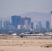 An aerial-ground level view of the Las Vegas skyline photographed from what appears to be a desert airfield or airport tarmac area. The iconic Stratosphere Tower (now known as STRAT Hotel) is prominently visible on the right side of the skyline, with numerous casino and hotel high-rises visible in the background. A military or civilian jet aircraft is visible on the runway/taxiway in the mid-ground.