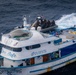 An aerial photograph shows a white and blue fishing vessel named 'Don Rufo' with registration number P.04.00875 being approached or boarded by armed personnel in tactical gear arriving via a rigid inflatable boat (RIB) flying a U.S. flag. The vessel's deck is loaded with yellow buoys, blue barrels, and other cargo. This appears to be a maritime law enforcement or interdiction operation.