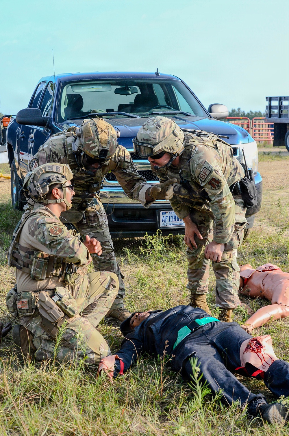 Three military personnel in OCP (Operational Camouflage Pattern) uniforms are conducting what appears to be a tactical combat casualty care (TCCC) training exercise outdoors. They are attending to training mannequins and a simulated casualty on the ground, with simulated wound moulage visible. A U.S. government vehicle with an Air Force license plate (AF11B0...) is visible in the background.