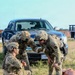 Three military personnel in OCP (Operational Camouflage Pattern) uniforms are conducting what appears to be a tactical combat casualty care (TCCC) training exercise outdoors. They are attending to training mannequins and a simulated casualty on the ground, with simulated wound moulage visible. A U.S. government vehicle with an Air Force license plate (AF11B0...) is visible in the background.
