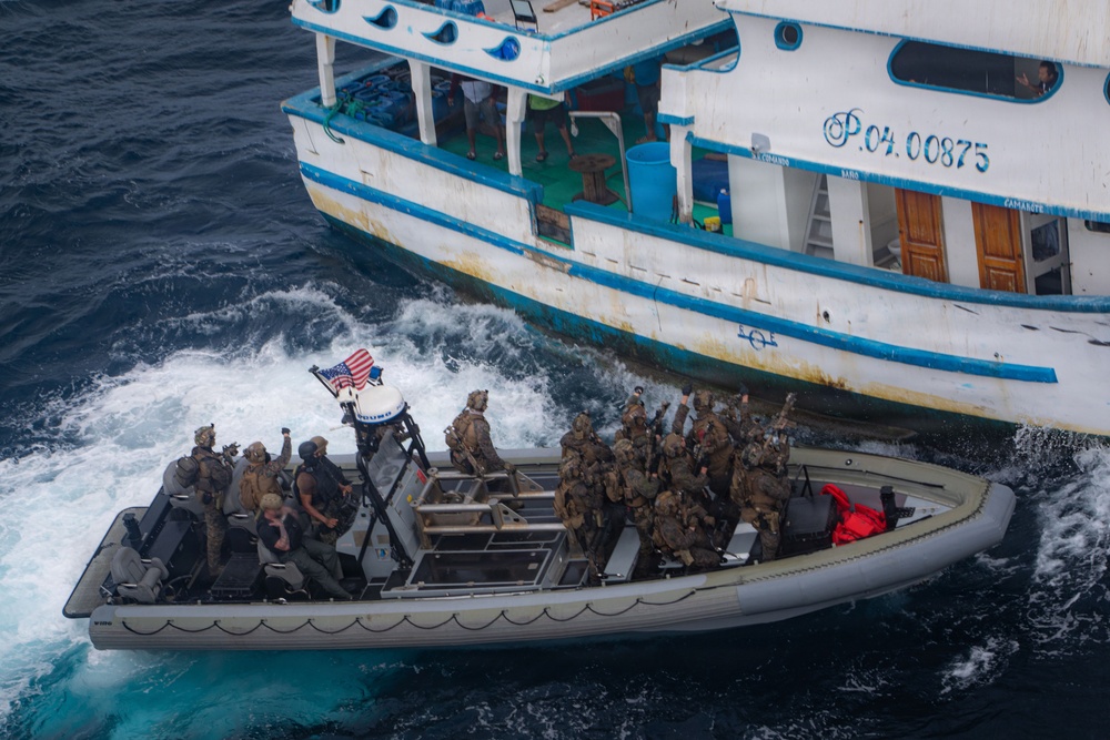 A rigid-hull inflatable boat (RHIB) carrying multiple armed personnel in military/tactical gear approaches a white civilian vessel marked 'P.04.00875' with blue trim. An American flag is visible on the RHIB, indicating a U.S. military or law enforcement maritime interdiction operation. The scene appears to be a vessel boarding or interception operation conducted on open ocean.