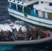 A rigid-hull inflatable boat (RHIB) carrying multiple armed personnel in military/tactical gear approaches a white civilian vessel marked 'P.04.00875' with blue trim. An American flag is visible on the RHIB, indicating a U.S. military or law enforcement maritime interdiction operation. The scene appears to be a vessel boarding or interception operation conducted on open ocean.