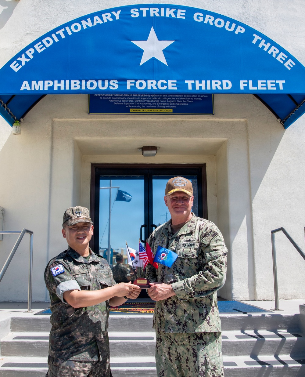 Two military personnel are posing in front of the Expeditionary Strike Group Three / Amphibious Force Third Fleet headquarters building. They are jointly holding a commemorative plaque or gift item featuring small flags, including what appear to be South Korean and American flags. The scene depicts a formal bilateral military exchange or visit between a Republic of Korea (ROK) military officer and a U.S. Navy officer.