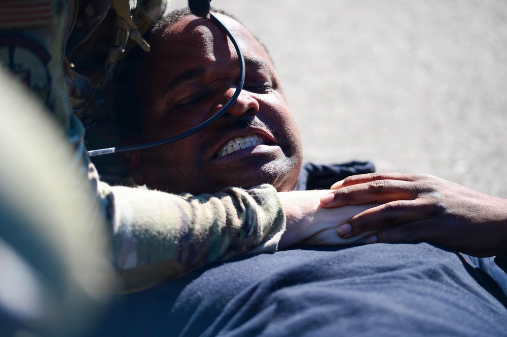 A person wearing military or tactical gear, including a helmet with attached equipment, is lying on a hard surface such as pavement. Another individual in camouflage clothing is leaning over them, with a hand placed on the person's chest or shoulder. The person on the ground appears to be grimacing or in distress, possibly during a training exercise or emergency scenario.