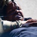A person wearing military or tactical gear, including a helmet with attached equipment, is lying on a hard surface such as pavement. Another individual in camouflage clothing is leaning over them, with a hand placed on the person's chest or shoulder. The person on the ground appears to be grimacing or in distress, possibly during a training exercise or emergency scenario.