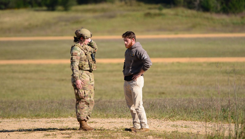 Two individuals are standing on a dirt path in an open field or training area. One person is wearing full military combat gear including camouflage uniform, body armor, and a helmet, while the other is dressed in civilian clothing. They appear to be engaged in a conversation or briefing.