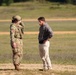 Two individuals are standing on a dirt path in an open field or training area. One person is wearing full military combat gear including camouflage uniform, body armor, and a helmet, while the other is dressed in civilian clothing. They appear to be engaged in a conversation or briefing.