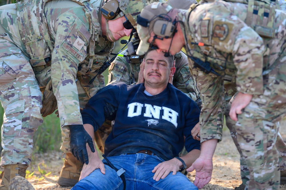 Three military personnel in MultiCam/OCP camouflage uniforms are attending to a civilian male wearing a UNH sweatshirt and jeans who is seated on the ground. The scene appears to be a training exercise or simulated casualty scenario conducted outdoors on a dirt surface. One soldier, identified by nametape as 'BURNETT,' is wearing tactical gloves and assisting alongside colleagues equipped with communications headsets.