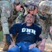 Three military personnel in MultiCam/OCP camouflage uniforms are attending to a civilian male wearing a UNH sweatshirt and jeans who is seated on the ground. The scene appears to be a training exercise or simulated casualty scenario conducted outdoors on a dirt surface. One soldier, identified by nametape as 'BURNETT,' is wearing tactical gloves and assisting alongside colleagues equipped with communications headsets.