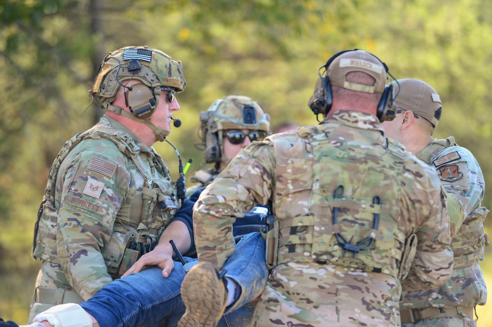 Multiple military personnel in MultiCam/OCP camouflage uniforms are conducting what appears to be a casualty evacuation or medical training exercise. They are carrying an individual wearing civilian clothing (blue jeans), suggesting a simulated rescue or MEDEVAC scenario. The setting is an outdoor wooded area with autumn foliage visible in the background.