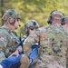 Multiple military personnel in MultiCam/OCP camouflage uniforms are conducting what appears to be a casualty evacuation or medical training exercise. They are carrying an individual wearing civilian clothing (blue jeans), suggesting a simulated rescue or MEDEVAC scenario. The setting is an outdoor wooded area with autumn foliage visible in the background.
