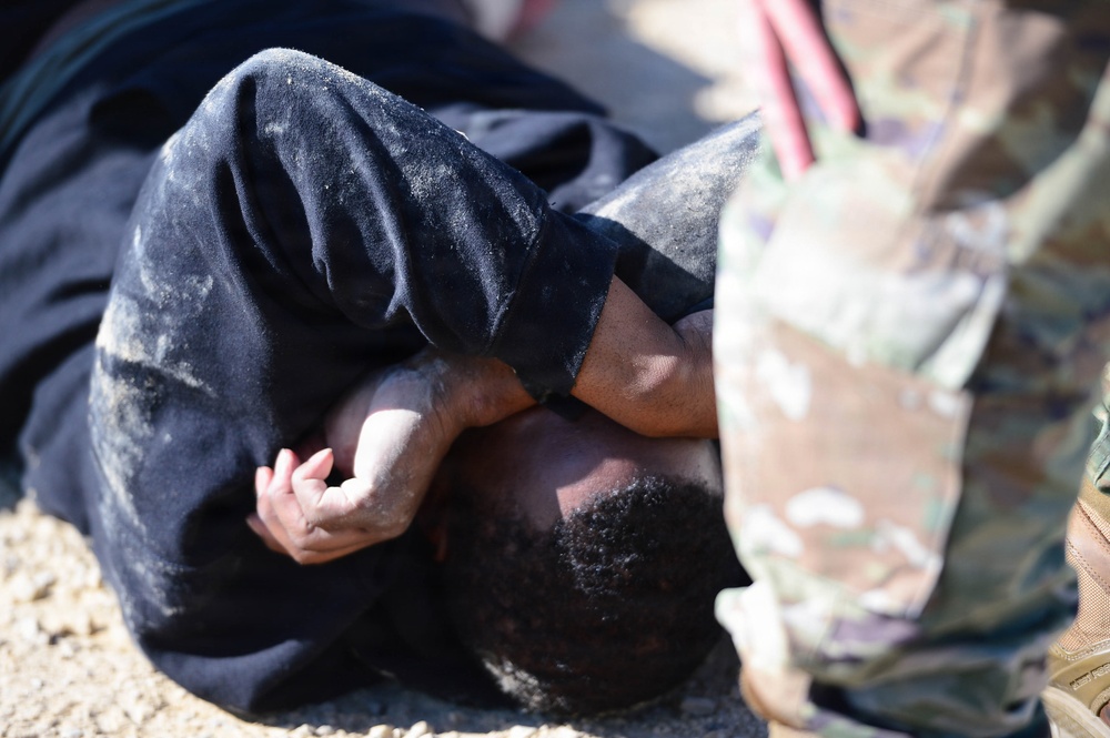 The image shows a person in a black, dust-covered garment lying on the ground in a curled or prone position, with hands clasped near their head. A second individual wearing military camouflage (MultiCam/OCP pattern) uniform and tan boots is visible in the foreground, partially out of focus. This appears consistent with a military training exercise, detainee handling drill, or combatives/ground fighting scenario.