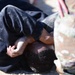 The image shows a person in a black, dust-covered garment lying on the ground in a curled or prone position, with hands clasped near their head. A second individual wearing military camouflage (MultiCam/OCP pattern) uniform and tan boots is visible in the foreground, partially out of focus. This appears consistent with a military training exercise, detainee handling drill, or combatives/ground fighting scenario.