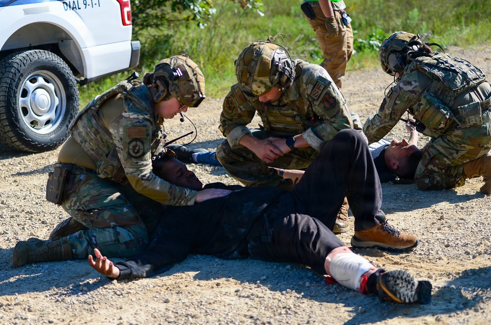 Military or law enforcement personnel in multicam/OCP camouflage uniforms are conducting what appears to be a tactical combat casualty care (TCCC) training exercise. Multiple personnel are attending to two individuals lying on a gravel surface, with visible simulated injuries including a bandaged leg wound with simulated blood. A white vehicle with a 'DIAL 9-1-1' decal is visible in the background, suggesting a domestic training environment.