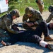 Military or law enforcement personnel in multicam/OCP camouflage uniforms are conducting what appears to be a tactical combat casualty care (TCCC) training exercise. Multiple personnel are attending to two individuals lying on a gravel surface, with visible simulated injuries including a bandaged leg wound with simulated blood. A white vehicle with a 'DIAL 9-1-1' decal is visible in the background, suggesting a domestic training environment.