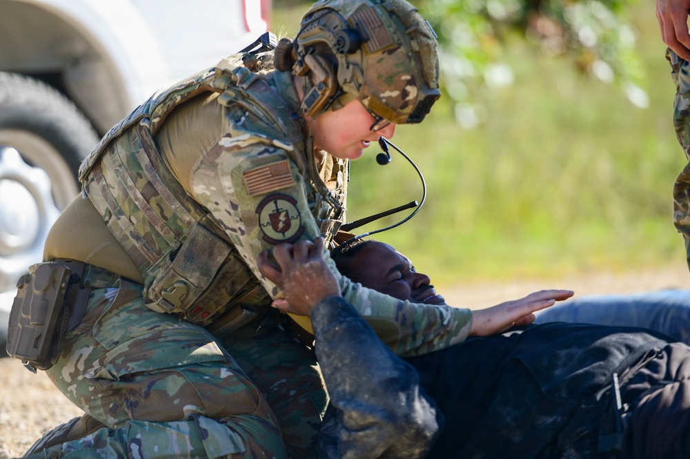 A U.S. military service member in full tactical gear, including a helmet with communications equipment and body armor, is kneeling over a person lying on the ground. The standing individual appears to be performing a medical assessment or casualty care procedure on the downed person. This appears to be a training exercise or tactical medical response scenario.