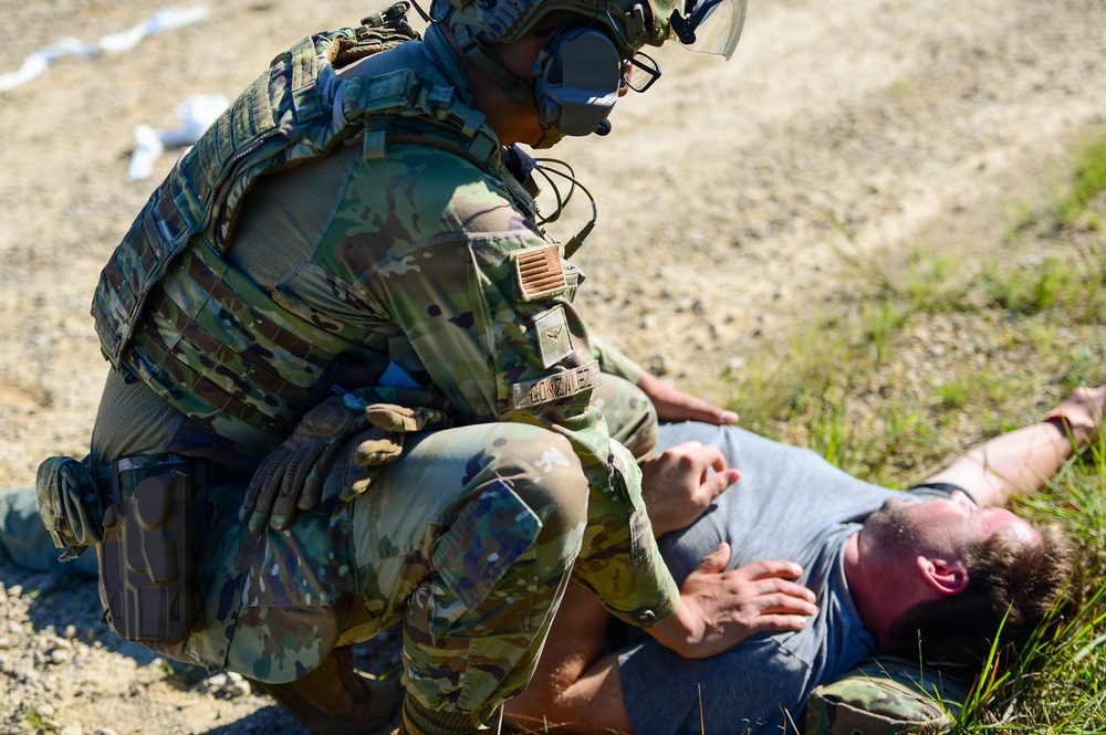 A military or law enforcement personnel in full tactical gear, identified by a name tape reading 'GONZALEZ,' is kneeling over a civilian male who is lying on the ground. The scene appears to depict a training exercise or tactical scenario involving casualty assistance or detainee control. The setting is an outdoor gravel and grass area in daylight conditions.