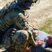 A military or law enforcement personnel in full tactical gear, identified by a name tape reading 'GONZALEZ,' is kneeling over a civilian male who is lying on the ground. The scene appears to depict a training exercise or tactical scenario involving casualty assistance or detainee control. The setting is an outdoor gravel and grass area in daylight conditions.