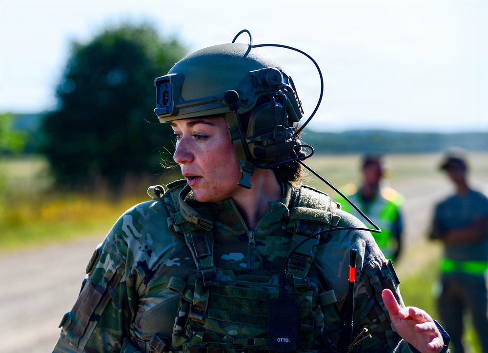 A female military service member in full tactical gear, including a combat helmet with communications equipment, is captured mid-conversation or briefing outdoors. She is wearing MultiCam/OCP camouflage uniform with a tactical vest and has a name tape reading 'OTTO' visible on her chest. Two blurred figures are visible in the background near what appears to be a tarmac or airfield.