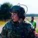 A female military service member in full tactical gear, including a combat helmet with communications equipment, is captured mid-conversation or briefing outdoors. She is wearing MultiCam/OCP camouflage uniform with a tactical vest and has a name tape reading 'OTTO' visible on her chest. Two blurred figures are visible in the background near what appears to be a tarmac or airfield.