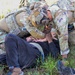 Two military personnel in full combat gear (OCP/MultiCam camouflage, tactical vests, helmets) are attending to a civilian individual lying on the ground in a grassy area. The scene appears to depict a tactical combat casualty care (TCCC) or medical assistance scenario, likely a training exercise. The personnel are leaning over the individual, appearing to assess or provide aid.