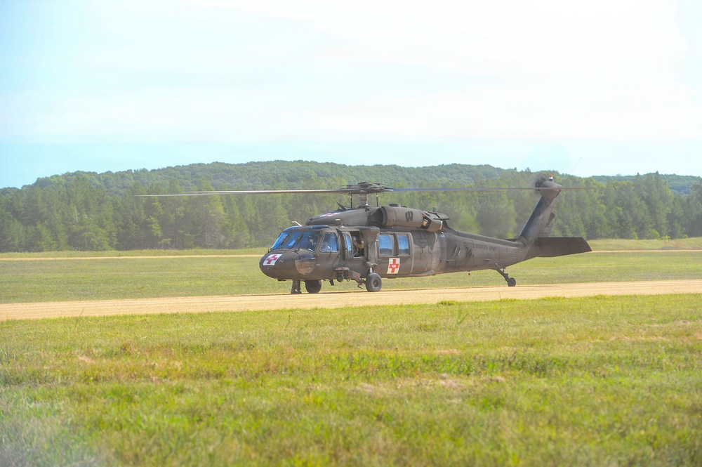 A U.S. Army UH-60 Black Hawk helicopter configured as a medevac (medical evacuation) aircraft is on or near a dirt landing strip in a grassy field. The helicopter displays red cross markings on its fuselage, indicating its medical role. The setting appears to be a military training area surrounded by dense tree lines.