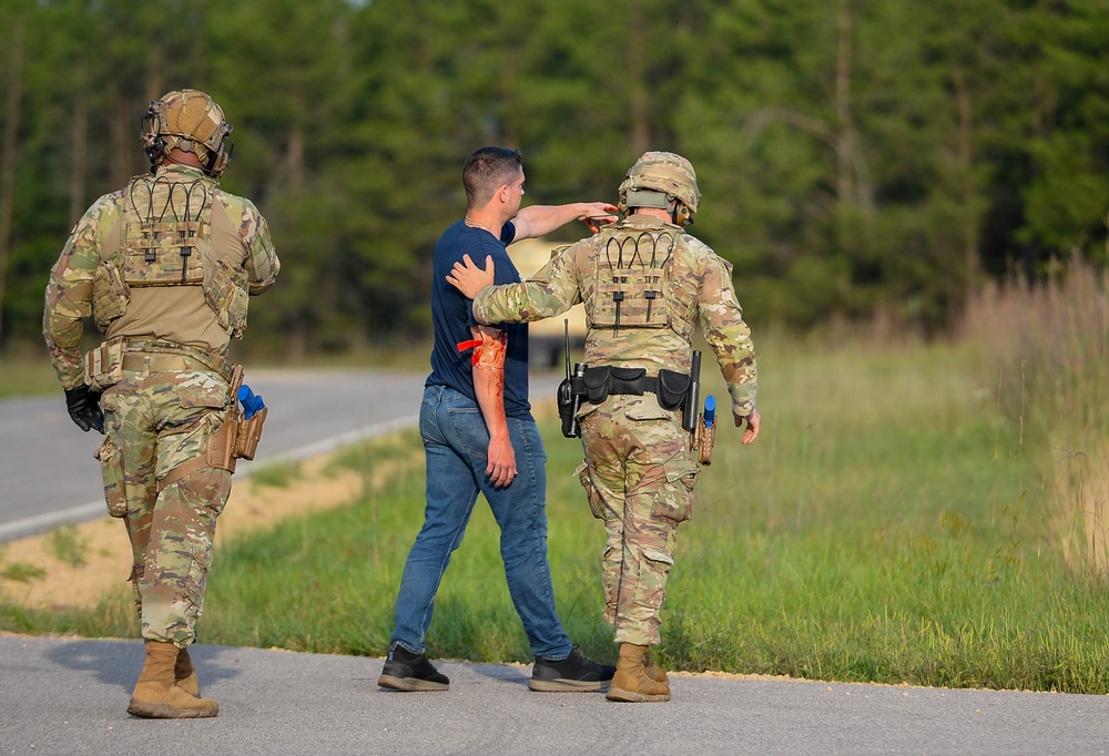 The image shows two soldiers in OCP (Operational Camouflage Pattern) military uniforms interacting with a civilian on a roadside near a wooded area. The civilian appears to have simulated injury markings (orange/red moulage) on their arm, suggesting this is a training exercise or drill. The soldiers are equipped with tactical gear, helmets, and radio equipment.