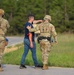The image shows two soldiers in OCP (Operational Camouflage Pattern) military uniforms interacting with a civilian on a roadside near a wooded area. The civilian appears to have simulated injury markings (orange/red moulage) on their arm, suggesting this is a training exercise or drill. The soldiers are equipped with tactical gear, helmets, and radio equipment.