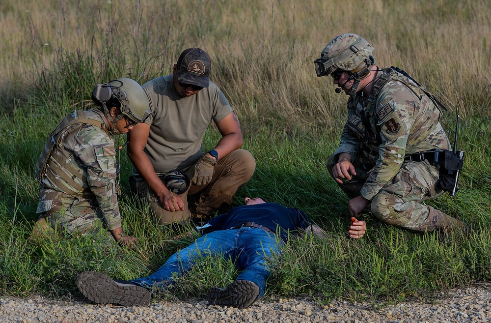 Three individuals in military/tactical attire are attending to a person lying on the ground in a grassy field area near a gravel path. Two personnel are wearing OCP (Operational Camouflage Pattern) uniforms with tactical gear, while a third wears civilian-style tactical clothing. This appears to be a military training exercise involving simulated casualty care or tactical combat casualty care (TCCC).