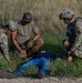 Three individuals in military/tactical attire are attending to a person lying on the ground in a grassy field area near a gravel path. Two personnel are wearing OCP (Operational Camouflage Pattern) uniforms with tactical gear, while a third wears civilian-style tactical clothing. This appears to be a military training exercise involving simulated casualty care or tactical combat casualty care (TCCC).