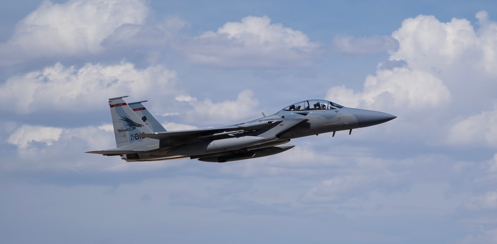 A twin-engine fighter jet, identifiable as an F-15 Eagle variant, is captured in level flight against a partly cloudy sky. The aircraft appears to be in a gray paint scheme with visible tail markings and is carrying external stores/weapons. The image is well-composed and sharp, suggesting a professional or military photography context.