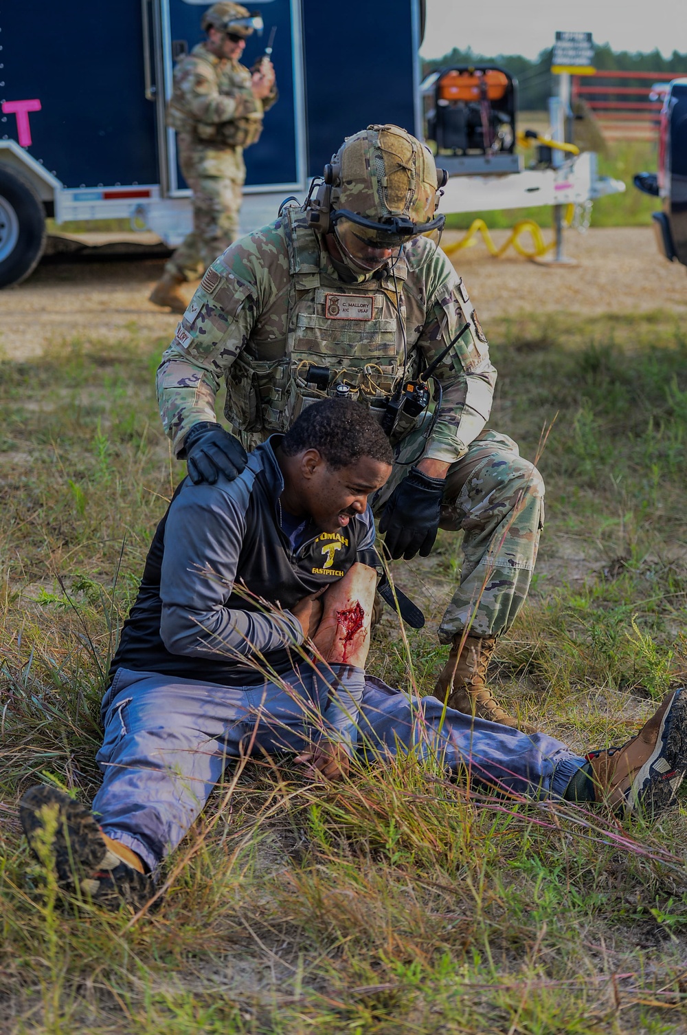 A U.S. Army soldier identified as C. Mallory is providing tactical combat casualty care to a civilian role-player during what appears to be a training exercise or disaster response drill. The civilian is seated on the ground displaying simulated wound makeup on their arm while the soldier kneels behind them providing assistance. A second soldier is visible in the background near a trailer, holding a radio.