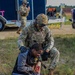 A U.S. Army soldier identified as C. Mallory is providing tactical combat casualty care to a civilian role-player during what appears to be a training exercise or disaster response drill. The civilian is seated on the ground displaying simulated wound makeup on their arm while the soldier kneels behind them providing assistance. A second soldier is visible in the background near a trailer, holding a radio.