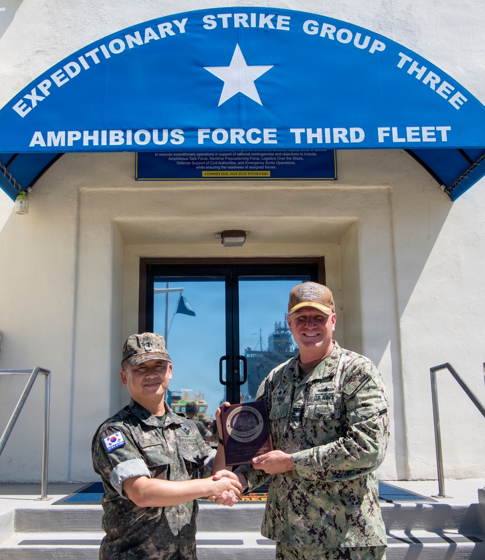 Two military officers are posing for a photo outside the headquarters of Expeditionary Strike Group Three, Amphibious Force Third Fleet. They are jointly holding a commemorative plaque while shaking hands, suggesting an official exchange or visit ceremony. One officer wears a Republic of Korea (ROK) military uniform and the other wears a U.S. Navy uniform.