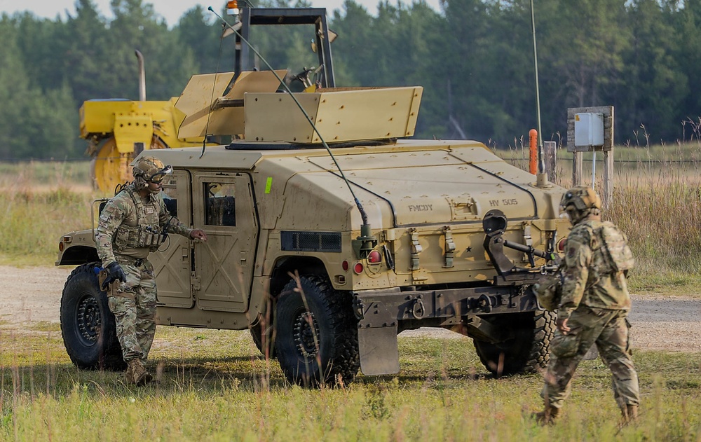 Two soldiers in camouflage uniforms (OCP/MultiCam pattern) are operating near a tan/sand-colored HMMWV (Humvee) military vehicle marked 'HV 505' and 'FMCOY' on a gravel path at what appears to be a training area. One soldier is positioned at the driver's side door while the other approaches from the rear. A yellow piece of heavy construction equipment is visible in the background.