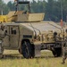 Two soldiers in camouflage uniforms (OCP/MultiCam pattern) are operating near a tan/sand-colored HMMWV (Humvee) military vehicle marked 'HV 505' and 'FMCOY' on a gravel path at what appears to be a training area. One soldier is positioned at the driver's side door while the other approaches from the rear. A yellow piece of heavy construction equipment is visible in the background.