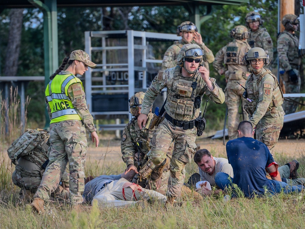 This image depicts a military training exercise involving U.S. Air Force Security Forces personnel responding to a simulated mass casualty or active threat scenario. Several uniformed personnel in OCP (Operational Camouflage Pattern) are attending to role-playing casualties on the ground, with one wearing a high-visibility 'POLICE' vest. The scene includes tactical gear, helmets, body armor, and radio communications consistent with a realistic training drill.