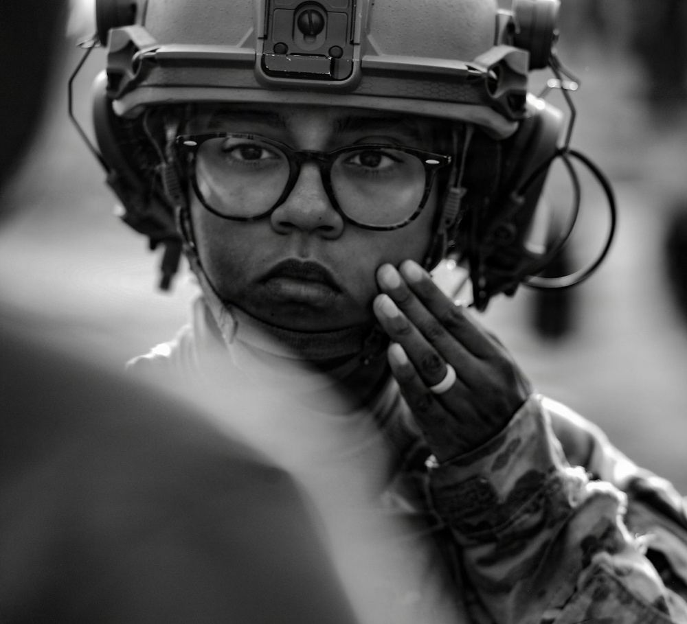 A black-and-white close-up portrait of a military or tactical personnel wearing a combat helmet with mounted accessories and communication headset. The individual is wearing eyeglasses and has one gloved hand raised near their face in a thoughtful or attentive pose. The image has a documentary or photojournalistic quality with strong focus on the subject's face.