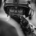 A black-and-white close-up portrait of a military or tactical personnel wearing a combat helmet with mounted accessories and communication headset. The individual is wearing eyeglasses and has one gloved hand raised near their face in a thoughtful or attentive pose. The image has a documentary or photojournalistic quality with strong focus on the subject's face.