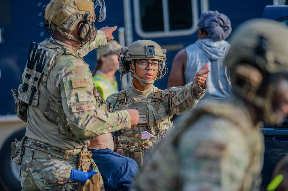 The image shows military personnel in full combat gear, including helmets and tactical vests, engaged in what appears to be a coordination or briefing discussion. Two soldiers are prominently visible with name tapes reading 'MALLORY' and 'HERNANDEZ', both wearing OCP (Operational Camouflage Pattern) uniforms. The setting appears to be an outdoor operational area with civilians and vehicles visible in the background.