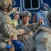 The image shows military personnel in full combat gear, including helmets and tactical vests, engaged in what appears to be a coordination or briefing discussion. Two soldiers are prominently visible with name tapes reading 'MALLORY' and 'HERNANDEZ', both wearing OCP (Operational Camouflage Pattern) uniforms. The setting appears to be an outdoor operational area with civilians and vehicles visible in the background.
