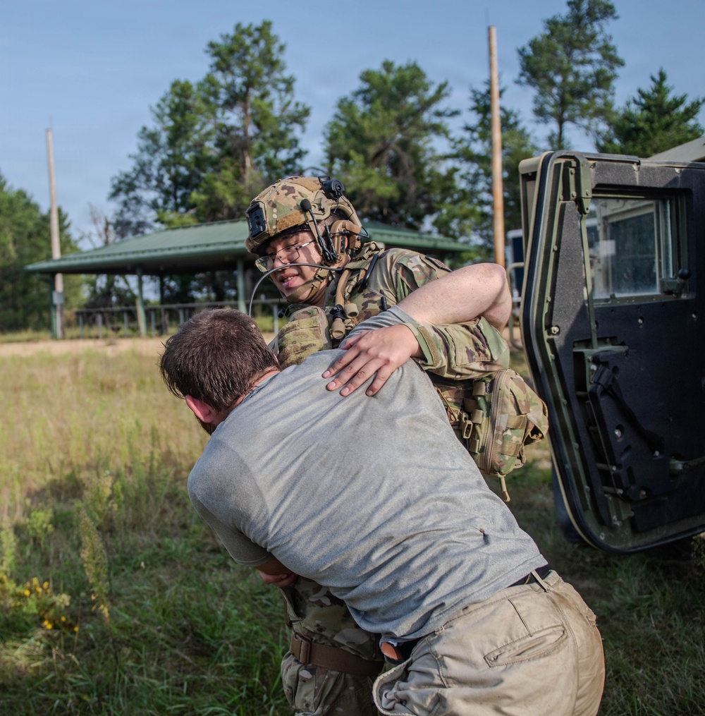 A soldier in full multicam combat gear, helmet, and communications headset is engaged in a physical grappling exercise with a civilian-clothed individual near a military HMMWV (Humvee). The scene appears to take place on a grassy field at a military training area, with trees and a small structure visible in the background. This appears to be a combatives or detainee-handling training scenario.