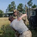 A soldier in full multicam combat gear, helmet, and communications headset is engaged in a physical grappling exercise with a civilian-clothed individual near a military HMMWV (Humvee). The scene appears to take place on a grassy field at a military training area, with trees and a small structure visible in the background. This appears to be a combatives or detainee-handling training scenario.