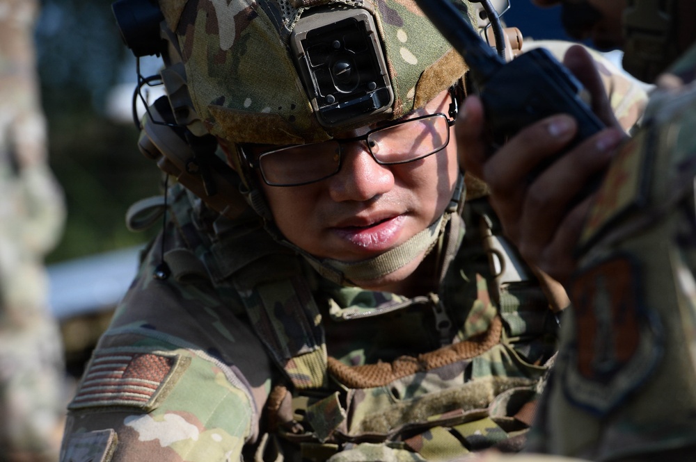 A U.S. Army soldier in full combat gear is closely examining a handheld radio or communication device. The soldier is wearing Operational Camouflage Pattern (OCP) uniform, a tactical helmet with a mounted device, ballistic glasses, and hearing protection. The image is a close-up shot that emphasizes the soldier's focused attention on the equipment.