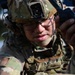 A U.S. Army soldier in full combat gear is closely examining a handheld radio or communication device. The soldier is wearing Operational Camouflage Pattern (OCP) uniform, a tactical helmet with a mounted device, ballistic glasses, and hearing protection. The image is a close-up shot that emphasizes the soldier's focused attention on the equipment.