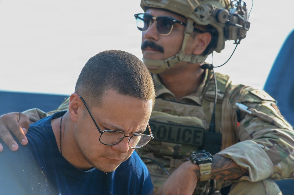 A law enforcement officer in tactical military-style gear with a 'POLICE' vest is detaining or escorting a civilian male. The officer is wearing a combat helmet with attachments, sunglasses, and camouflage uniform, and has his hand on the civilian's shoulder. The scene appears to be an outdoor law enforcement operation.