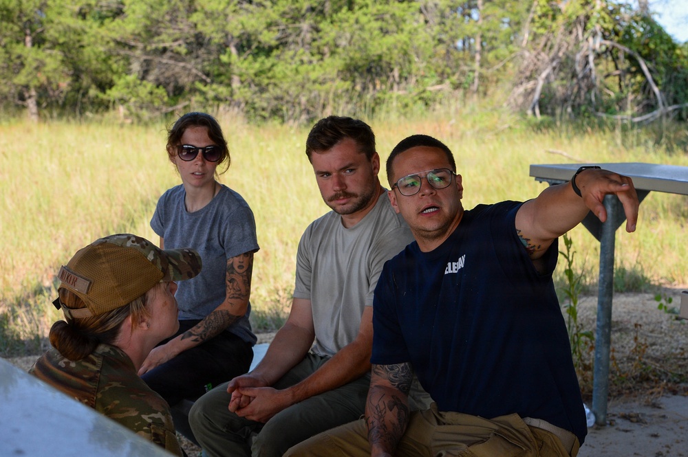 Four individuals are gathered outdoors in what appears to be a briefing or instructional setting near a metal table or bench. One person wearing a navy blue shirt and glasses is gesturing and appears to be explaining or pointing something out to the group. The setting includes tall grass and trees in the background, suggesting a rural or training area.